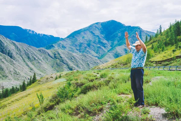 Joyous man near highway among giant mountains. Cheerful traveler on hill among rich vegetation. Mountain tourism. Journey in highland. Amazing mountainous landscape of majestic nature. Active rest.