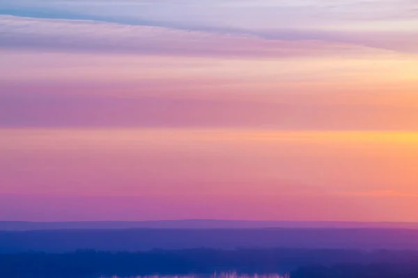 Varicolored striped surreal sky with shades of blue, cyan, pink, purple, magenta colors with cobalt land and lake. Horizontal lines of smooth clouds. Atmospheric image of tender sky, land and river.