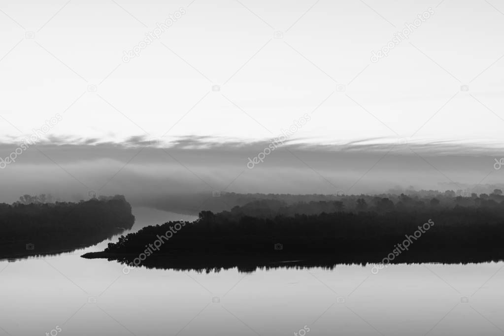 Silhouette of river shore with forest under early thick fog in grayscale. Mystical smooth water. Riverbank under cloudy sky. Morning atmospheric minimalistic landscape of majestic nature monochrome.