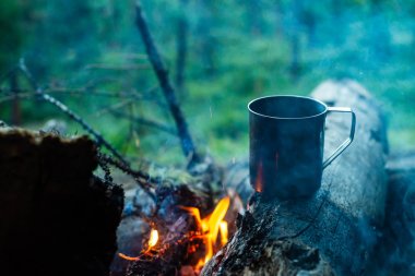 Metal kupadaki çay şenlik ateşinde taşüzerinde ısınır. Doğada sıcak içecek. Açık havada çay içmek. Gün batımında kamp. Alacakaranlıkta romantik sıcak atmosfer açık . Aktif dinlenme.