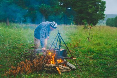 Turist ateşi yakSa. Gezgin kampta ateş yakıyordu. Adam kamp ateşine kuru dal koyar. Aktif açık hava rekreasyon. Vahşi doğaya. Gün batımında kamp. Alacakaranlıkta atmosferik sıcak. Aktif dinlenme.