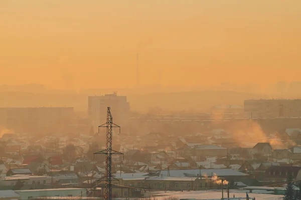 Power lines in city on dawn. Silhouettes of urban buildings among smog on sunrise. Cables of high voltage on warm orange yellow sky. Power industry at sunset. City power supply. Mist urban background.