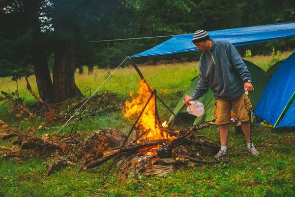 Tourist kindles fire. Traveler is lighting fire in camp. Man by campfire. Active outdoor recreation into wild. Camping in dusk. Atmospheric warm in twilight. Active rest. Hot tea in kettle on bonfire.