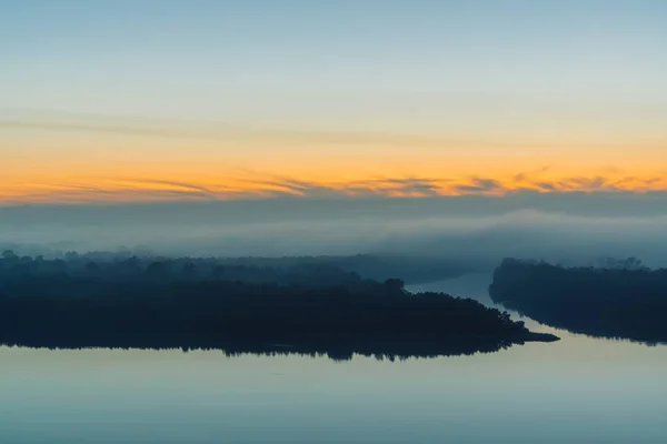 Broad river flows along shore with forest under thick fog. Early blue sky reflected in water. Yellow glow in picturesque predawn sky. Colorful morning mystical atmospheric landscape of majestic nature