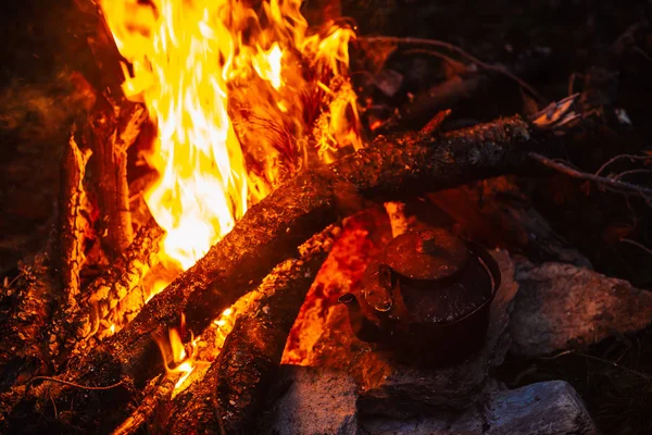 Boiling of tea in kettle on bonfire with large firewood. Tea drinking in open air. Active outdoor recreation. Camping in dusk. Romantic warm atmosphere in twilight on nature. Active rest.