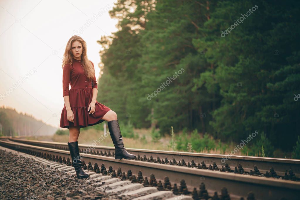 Beautiful sad thoughtful girl with curly natural hair on nature in forest on railway. Dreamer lady in burgundy dress walk on railroad. Depressed girl on rails at dawn. Sun in hair in autumn. Bad mood.