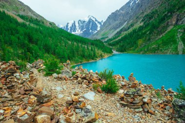 Buzul ve dağ gölünün arka planında taşlı tepeüzerinde taş dengeleme taşları Piramitleri. Yaylaların zengin bitki örtüsü. Dağ yamacında kozalaklı orman. Harika dağlar. İnanılmaz canlı yeşil manzara.