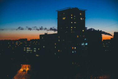 Sıcak gün batımı gökyüzünün arka planında aydınlık pencerelere sahip inanılmaz bina siluetleri. Şafakta şehrin üstündeki borudan duman. Alacakaranlıkta parlayan pencereler. Minimalist şehir manzarası. Harika kentsel gündoğumu.