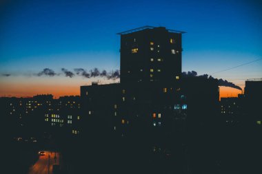 Sıcak gün batımı gökyüzünün arka planında aydınlık pencerelere sahip inanılmaz bina siluetleri. Şafakta şehrin üstündeki borudan duman. Alacakaranlıkta parlayan pencereler. Minimalist şehir manzarası. Harika kentsel gündoğumu.