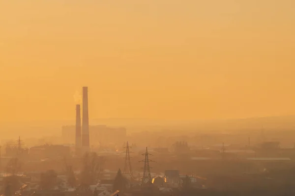 Smog among silhouettes of buildings on sunrise. Smokestack in dawn sky. Environmental pollution on sunset. Harmful fumes from stack above city. Mist urban background with warm orange yellow sky.