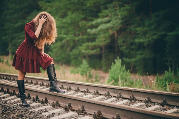 Beautiful sad girl is hiding face by hair. Moody lady in burgundy dress in forest on railway. Depressed lonely girl on railroad at dawn. Sun in curly natural hair in autumn. Bad mood. Offended girl.