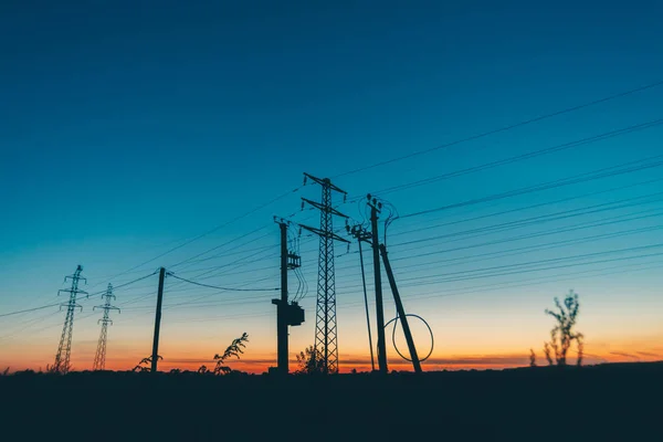 日の出の背景にフィールドの電力線 夜明けにワイヤーを持つポールのシルエット 暖かいオレンジ色の青空に高電圧のケーブル 日没時の電力産業 絵のように鮮やかな空に多くのケーブル — ストック写真