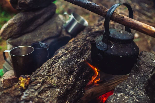 Boiling of tea in kettle on bonfire with large firewood. Tea drinking in open air. Active outdoor recreation. Camping in dusk. Romantic warm atmosphere in twilight on nature. Active rest. Metal mug.