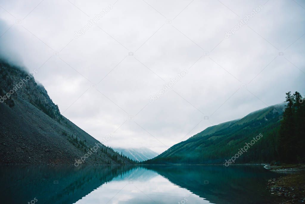 Giant cloud above rocky mountainside with trees in fog. Amazing mountain lake. Mountain range under cloudy sky. Wonderful rocks in mist. Morning landscape of highland nature. Low clouds in mountains.