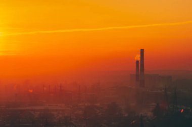 Smog among silhouettes of buildings on sunrise. Smokestack in dawn sky. Environmental pollution on sunset. Harmful fumes from stack above city. Mist urban background with warm orange yellow sky. clipart