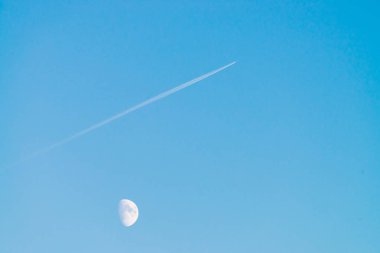 Berrak mavi gün gökyüzünde ay üzerinde jet yoğuşma izi. Minimalist mavi arka plan. Uçak çapraz olarak uçar. Uçak hava sahasında uçuyor. Gökyüzündeki beyaz ay.