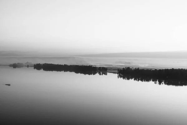 Broad river flows along diagonal shore with silhouette of forest and thick fog in grayscale. Tree drifts with flow. Minimalistic monochrome landscape of majestic nature. Morning milky atmosphere.