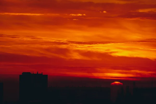 Sun rises over horizon. Fiery sunny circle. Cityscape with vivid red dawn. Amazing warm dramatic cloudy sky above dark silhouettes of city building roofs. Atmospheric background of sunrise. Copy space