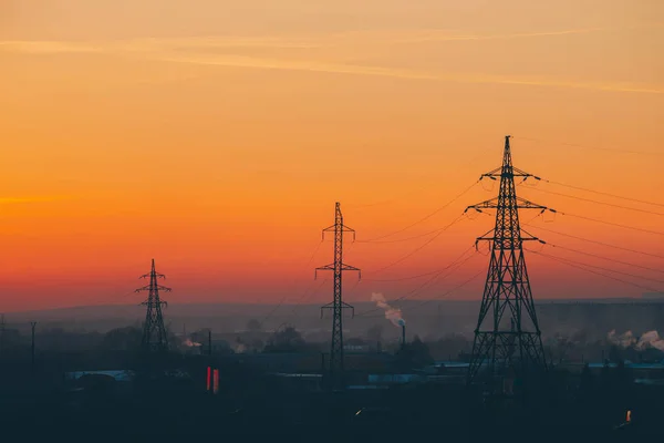 Şafakta şehirde elektrik hatları. Gün doğumunda sis arasında kentsel binaların siluetleri. Sıcak turuncu sarı gökyüzünde yüksek voltaj kabloları. Gün batımında güç endüstrisi. Şehir güç kaynağı. Sis kentsel arka plan.
