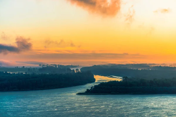 Azure river flows along shore with forest under fog. Channel of river flows around island. Orange glow in dawn sky. Colorful morning mystical atmospheric landscape of majestic nature.