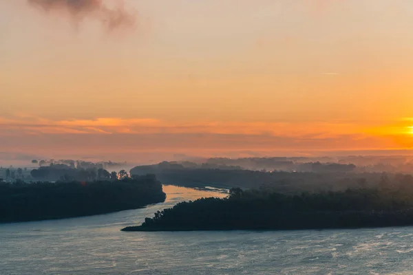 River flows along shore with forest under fog. Channel of river flows around island. Orange glow in dawn sky reflected on water. Colorful morning mystical atmospheric landscape of majestic nature.