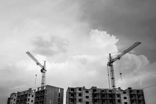 Cranes above unfinished multistorey panel building on background of cloudy sky with copy space in grayscale. Process of construction of apartment building in overcast weather close up in monochrome.