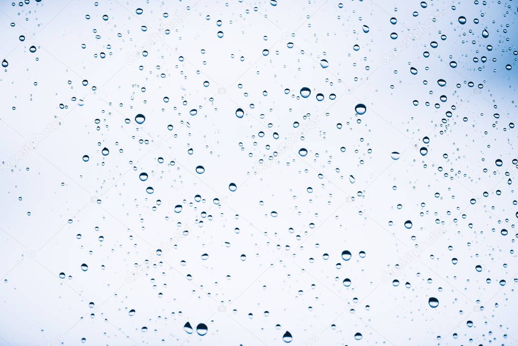 Dirty window glass with drops of rain. Atmospheric blue light background with raindrops. Droplets and stains close up. Detailed transparent texture in macro with copy space. Rainy weather.