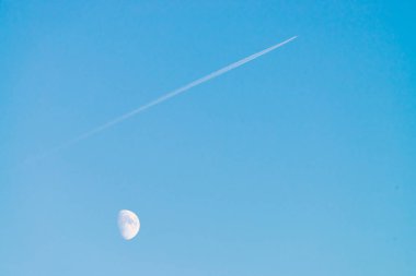 Berrak mavi gün gökyüzünde ay üzerinde jet yoğuşma izi. Minimalist mavi arka plan. Uçak çapraz olarak uçar. Uçak hava sahasında uçuyor. Gökyüzündeki beyaz ay.