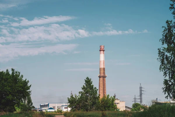 Giant smoke stack in industrial area behind trees. Industrial structure with big pipe of brown brick under blue sky. Technology and nature. Pollution of environment. Vegetation in industry background.