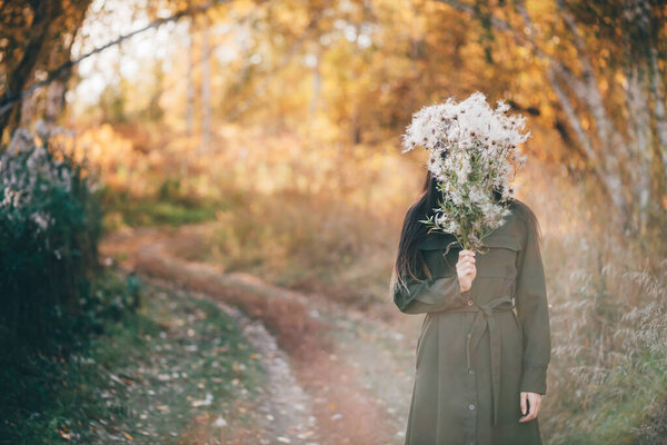 Dreamy beautiful girl in golden sunlight on bokeh background with yellow leaves. Inspired girl enjoys sunset in autumn forest. Girl hiding her face behind thistle flowers bouquet among autumn foliage.
