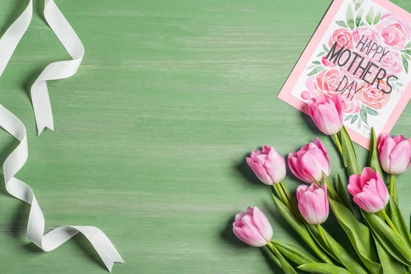Vista elevada de fita branca, buquê de tulipas rosa e cartão com letras dia feliz mães no fundo verde — Fotografia de Stock