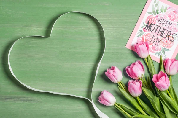 Vista elevada do símbolo do coração feito de fita, buquê de tulipas e cartão com letras feliz dia das mães no fundo verde — Fotografia de Stock