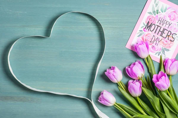 Top view of heart symbol made from ribbon, bouquet of tulips and card with lettering happy mothers day on blue background — Stock Photo