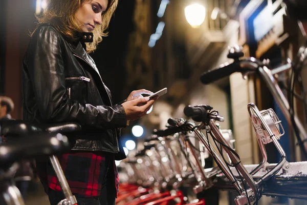Young girl in black leather jacket using in hands screen smartphone on background bokeh light in night city street, hipster biking and riding to job by ecology  bicycle, lifestyle online wifi internet concept
