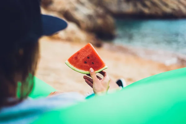 young girl hipster relax on beach coast and holds in her hand a slice of red fresh fruit watermelon on blue sea background, woman on seaside nature eating sweet healthy food, vacation in summer concept