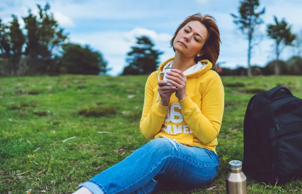 Happy girl with eyes closed holding in hands cup of hot tea on green grass in outdoors nature park, beautiful woman hipster enjoy drinking cup of coffee, lifestyle relax recreation meditation concept