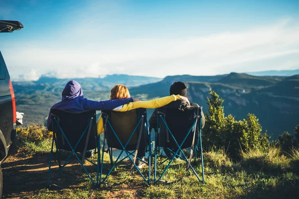 three friends sit in camping chairs on top of a mountain, travelers enjoy nature and cuddle, tourists look into distance on background of panoramic landscape, weekend concept