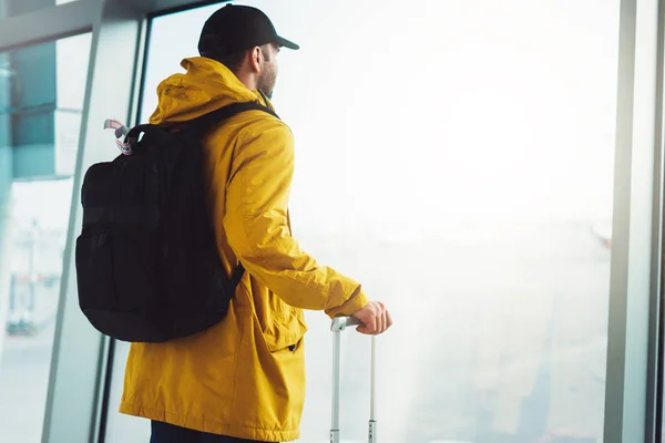 traveler tourist with yellow backpack is standing at airport on background large window, man in bright jacket waiting in departure lounge area hall of  airport lobby terminal, vacation trip concept, empty space mockup