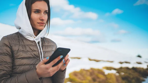 girl hold in hands mobile phone, tourist planning trip in snow mountain on blue sky background, person type message on smartphone in landscape trip, hipster enjoy winter nature, lifestyle holiday concept
