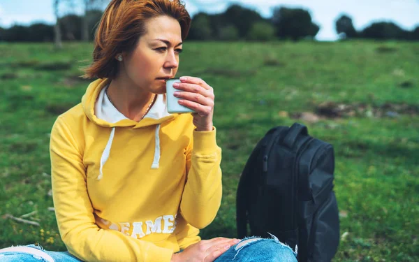 Happy smile girl holding in hands cup of hot tea on green grass in outdoors nature park, beautiful woman hipster enjoy drinking cup of coffee, lifestyle relax recreation meditation concept