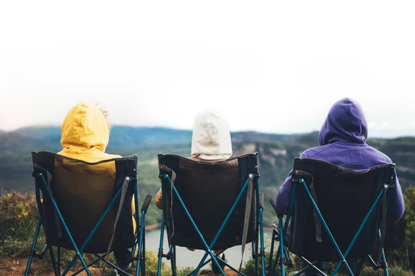 three friends sit in camping chairs on top of a mountain, travelers enjoy nature and cuddle, tourists look into distance on background of panoramic landscape, weekend concept