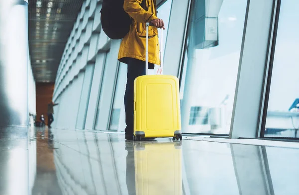 traveler in bright jacket with yellow suitcase backpack at airport on background large window blue sky, passenger waiting flight in departure lounge area, hall of airport lobby terminal, vacation trip concept, empty space mockup