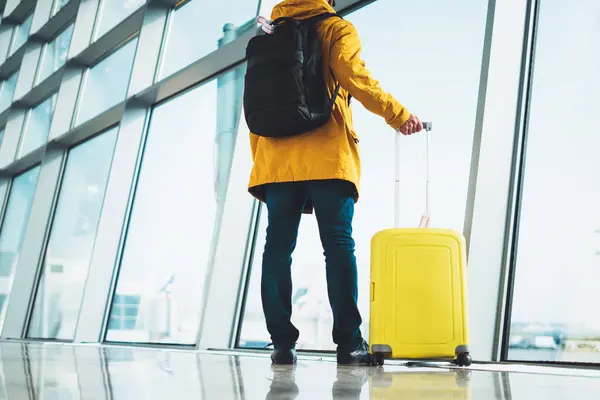 traveler in bright jacket with yellow suitcase backpack at airport on background large window blue sky, passenger waiting flight in departure hall of lobby terminal lounge area, vacation trip concept, empty space mockup