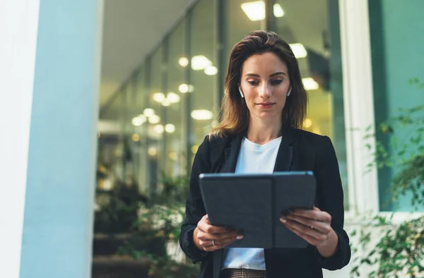 Pensive female student of faculty of law checking mail and reading notification with financial news on tablet device standing outdoors near evening neon lights