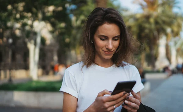 girl smiles at good news with her smartphone. Portrait girl in a white t-shirt walking in a Park with palm trees and using Internet technology in sell phone