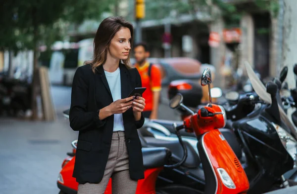 pretty girl in business clothes holds  smartphone background of rent mopeds in city of summer street outdoors, active tourist using mobile phone in female hands and plans in online app device walk around busy city scooter