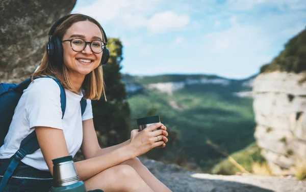 portrait girl smile travel in mountain drinking coffee from thermos enjoying summer trip, fun female hipster tourist with glasses relaxing in nature vacation listening music with headphones, copy space for text