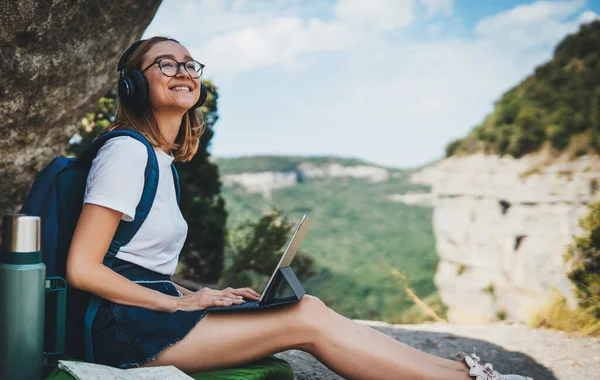young girl with glasses and backpack enjoying summer vacation in mount listen music in headphones and laptop in nature, fun traveler woman planning hiking walk looks at online map on tablet device