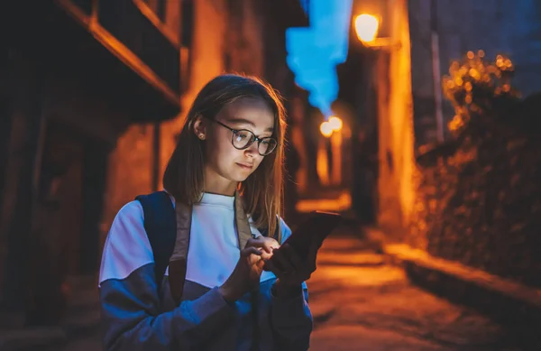 portrait tourist girl using mobile phone with eyeglasses reflection of screen light backdrop of street old historical city at night, young woman traveler plans walk through architectural sights of old town looks at online map in smartphone