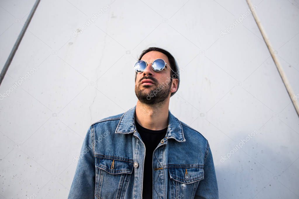 portrait of handsome bearded man posing in denim jacket and sunglasses near wall